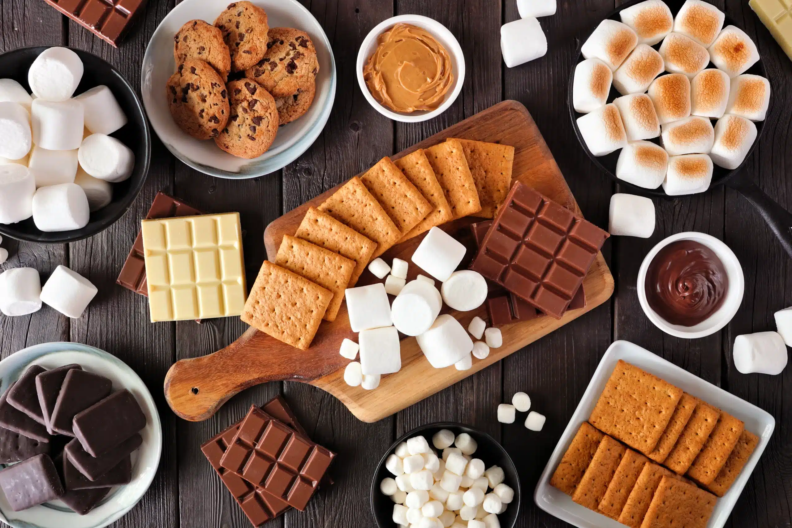 A table covered with plates and bowls with ingredients for s'mores: bars of chocolate, graham crackers, and marshmallows